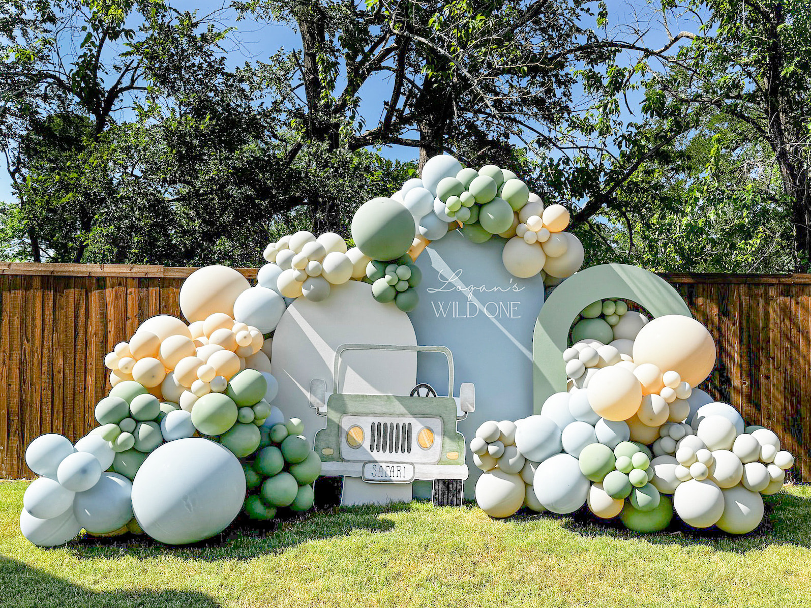 A colorful and whimsical balloon arch adorns the entrance to a grassy outdoor area, with a vintage-style truck parked in the background amidst lush greenery.