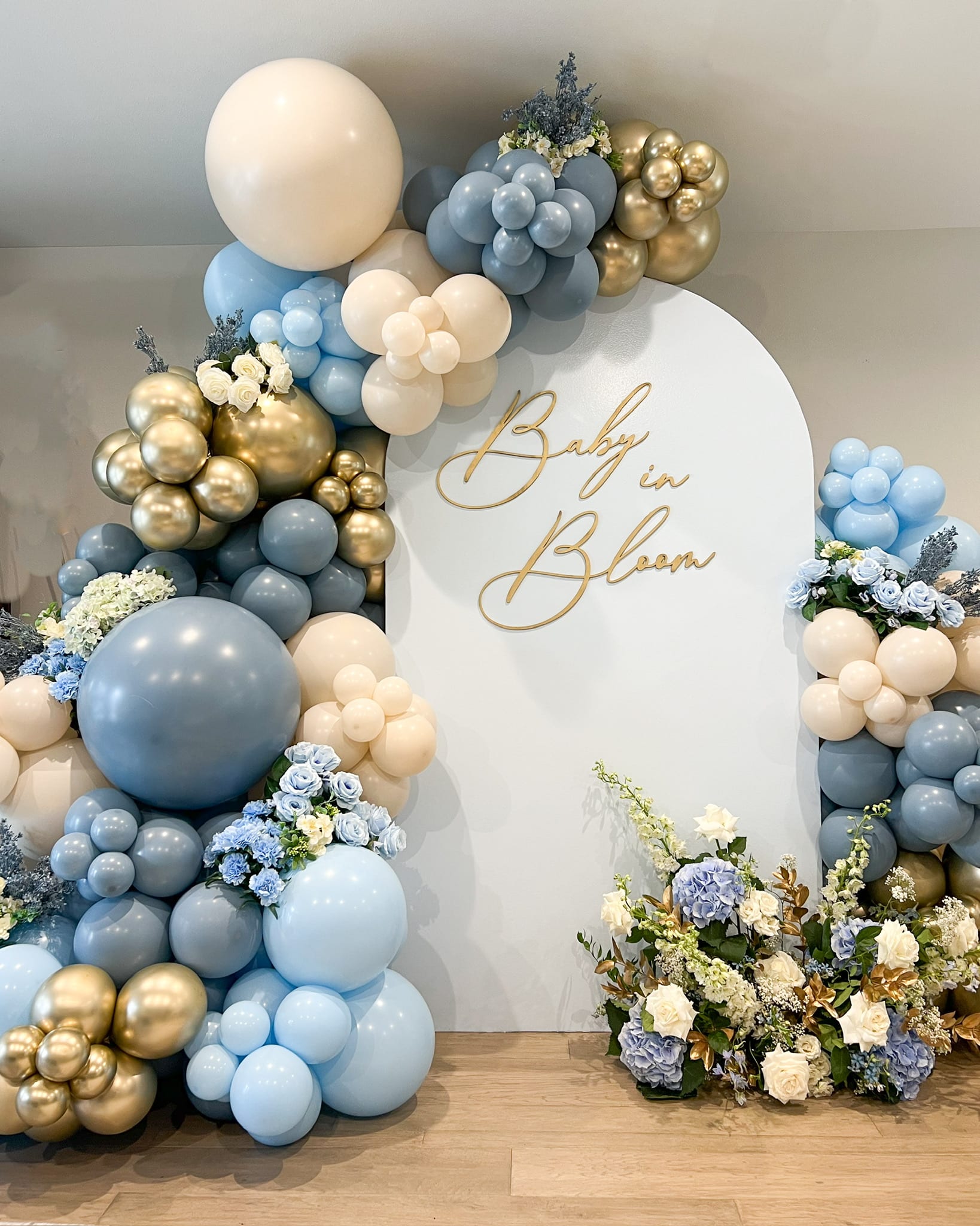 A decorative arrangement of white, blue, and gold balloons, along with a floral display, forms the centerpiece of the image, which appears to be a baby shower or gender reveal celebration.