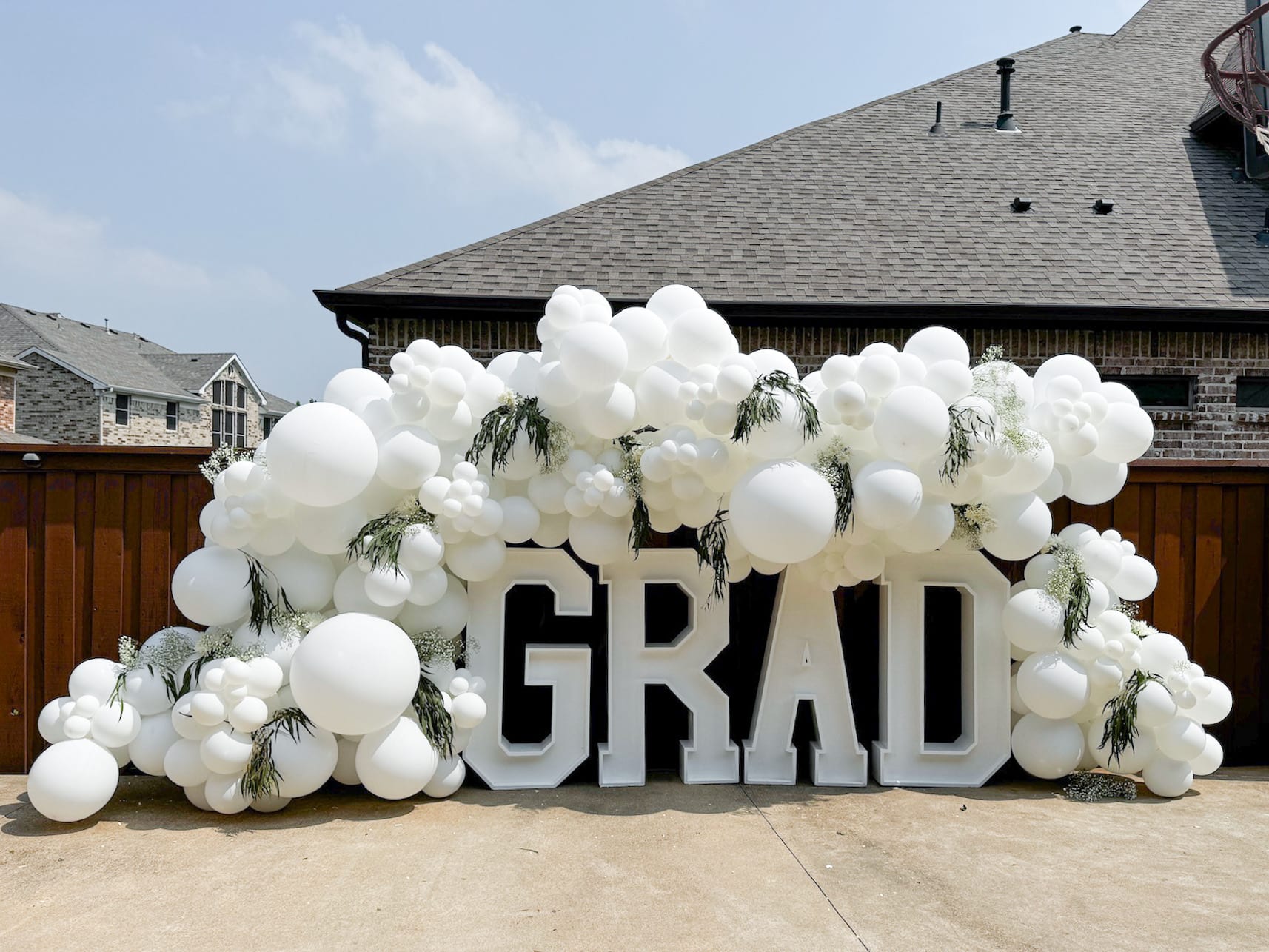 A large display of white balloons arranged in the shape of the word "GRAD" stands in the foreground, with a wooden building visible in the background.