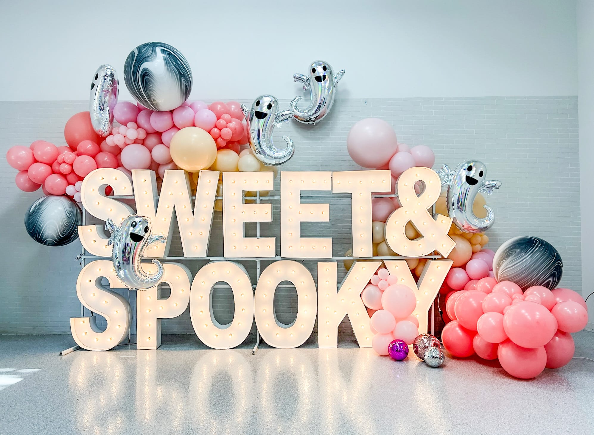The image features the text "SWEET & SPOOK" prominently displayed in the foreground, surrounded by various colorful balloons in shades of pink, yellow, and black. The background appears to be a plain, light-colored surface, creating a minimalist and visually striking composition.