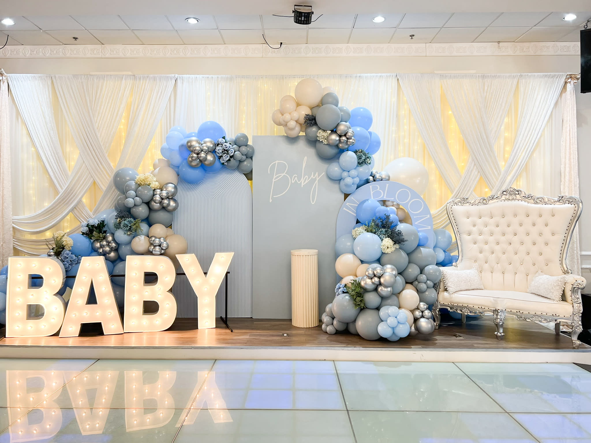 The image depicts an elaborate baby shower setup, with large "BABY" letters, a tufted armchair, and a decorative balloon arch in shades of blue and gray, creating a warm and celebratory atmosphere.