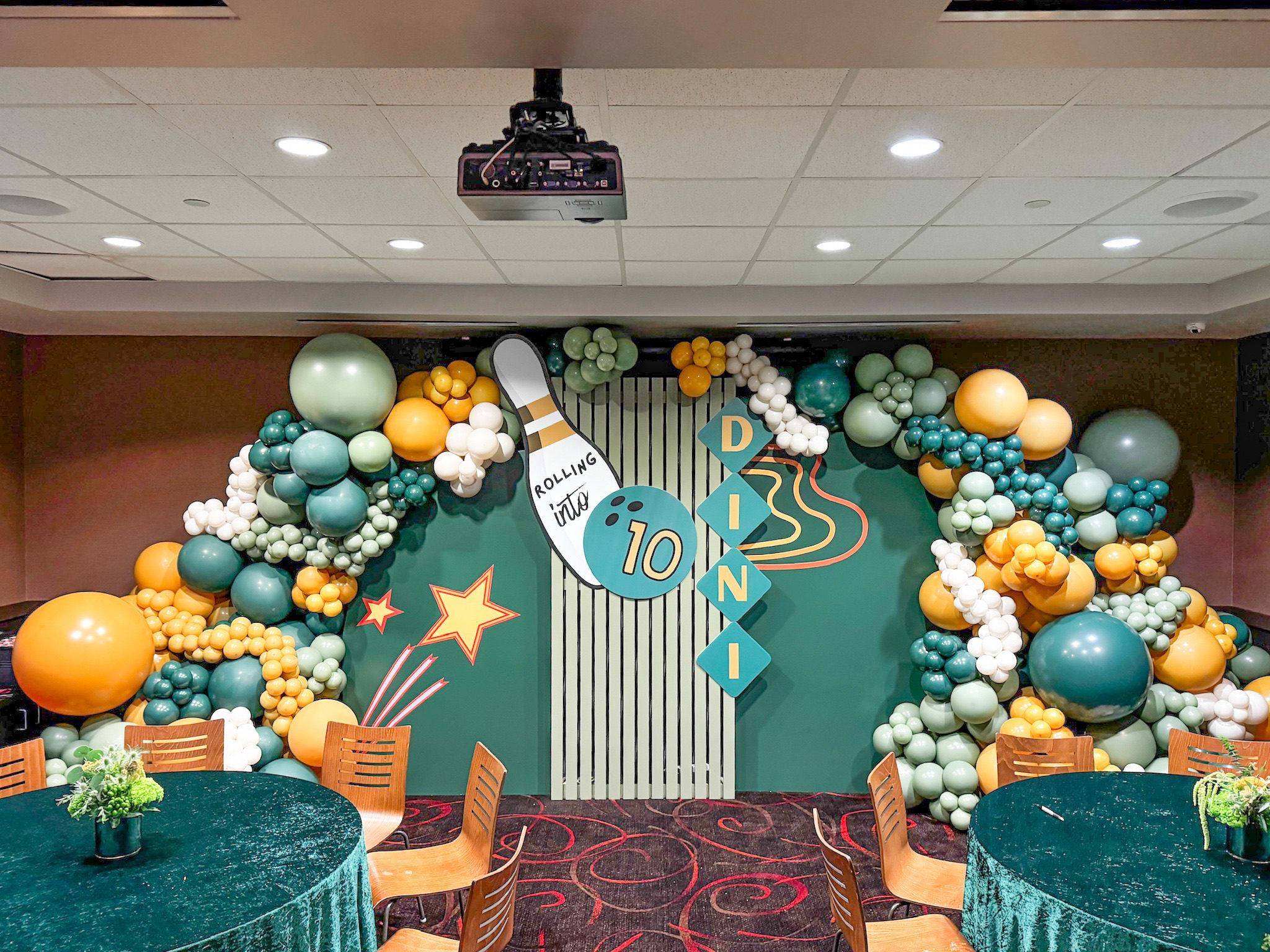 A colorful and whimsical balloon arch adorns the entrance, with a variety of balloons in shades of green, orange, and blue. The background features a wooden-paneled wall and a projector screen, creating a festive and celebratory atmosphere.