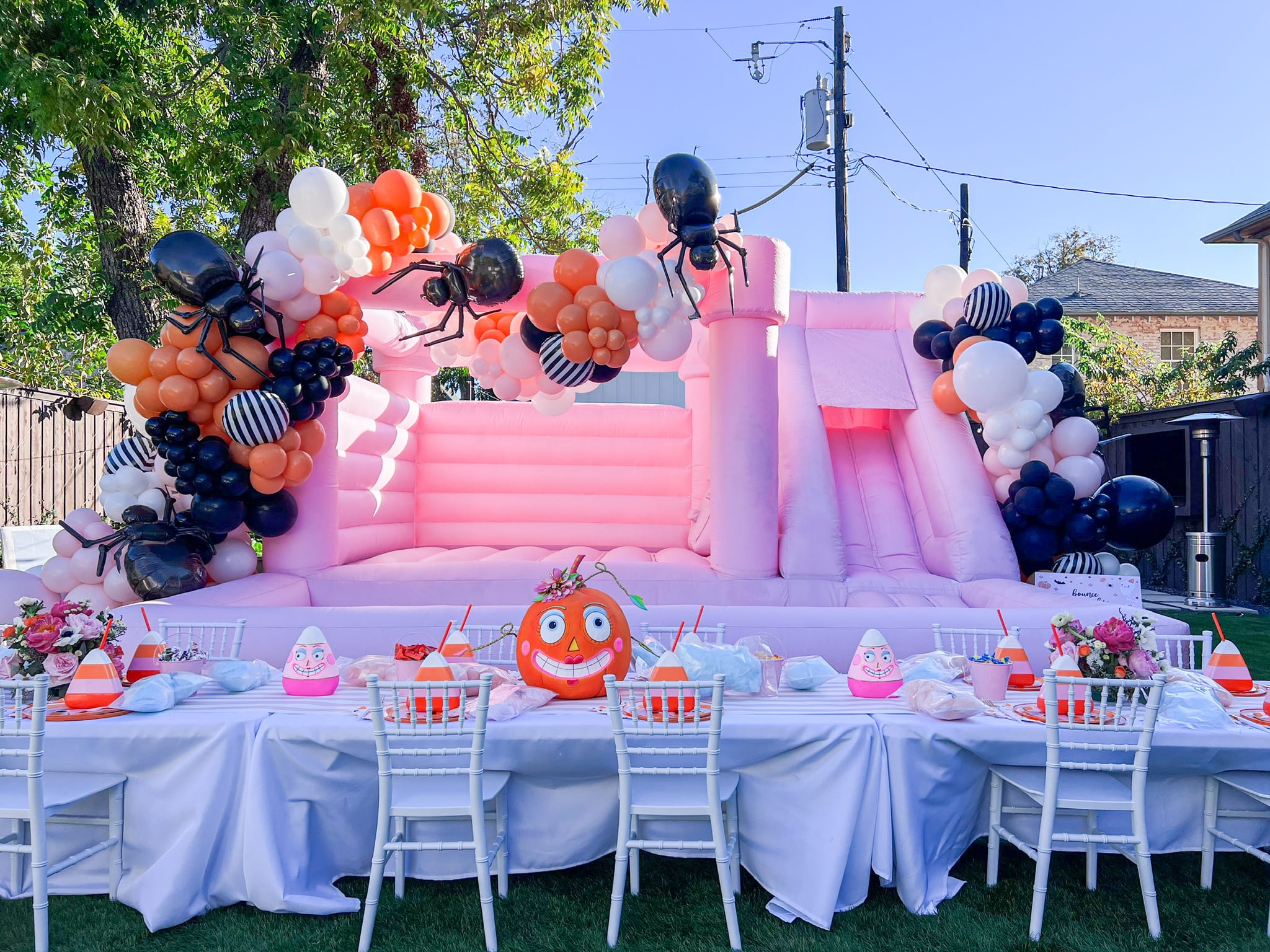 A vibrant and whimsical outdoor party setup, featuring a large inflatable structure in pink, surrounded by colorful balloons, tables with place settings, and a cheerful character decoration.