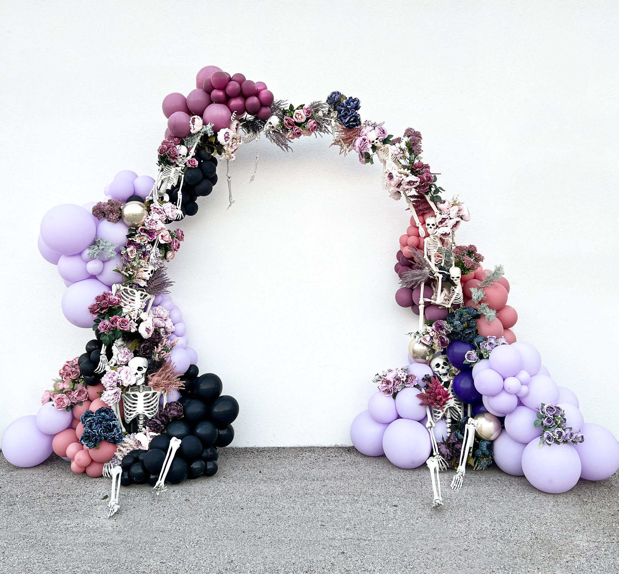 A decorative arch made of colorful balloons, flowers, and skeletal figures stands against a plain gray background.