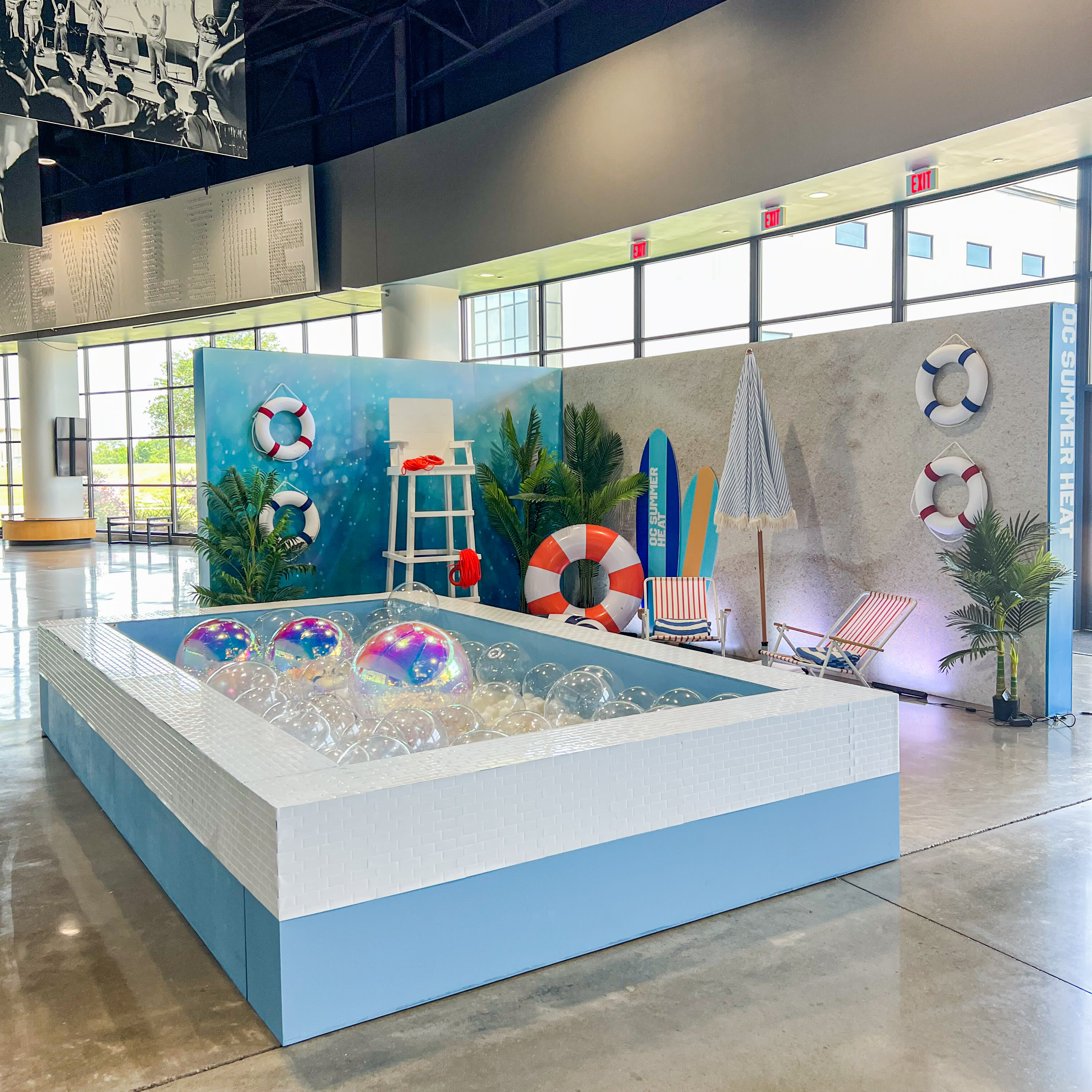 A large indoor pool surrounded by colorful beach-themed decor, including surfboards, life preservers, and tropical plants, set against a backdrop of windows overlooking an outdoor area.