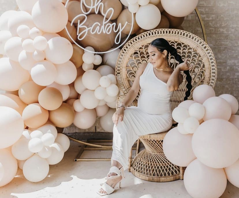 a woman in a white dress sitting on a wicker chair in front of a backdrop of white and pink balloons