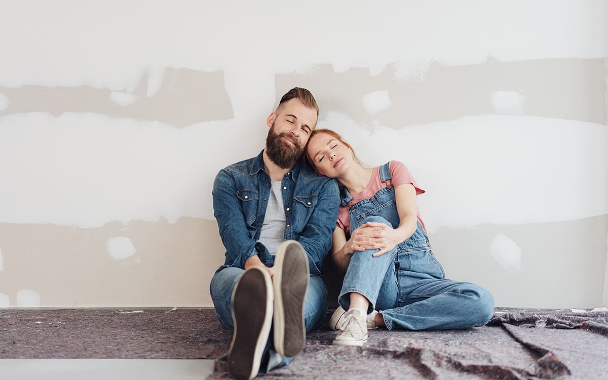 Ein Paar sitzt auf dem Boden eines Raumes, der renoviert wird; sie lehnen ihre Köpfe aneinander und lächeln mit geschlossenen Augen.