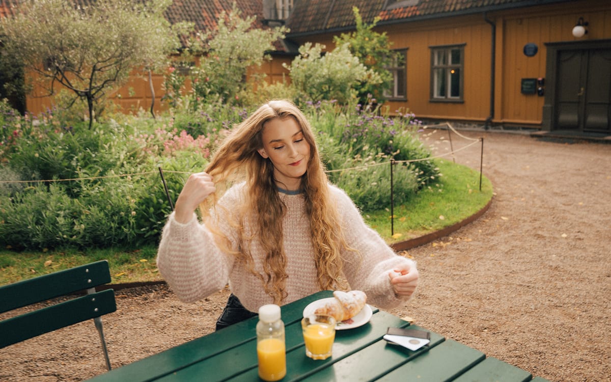 Kvinne med langt, bølget hår sitter på en kafé med croissant og appelsinjuice