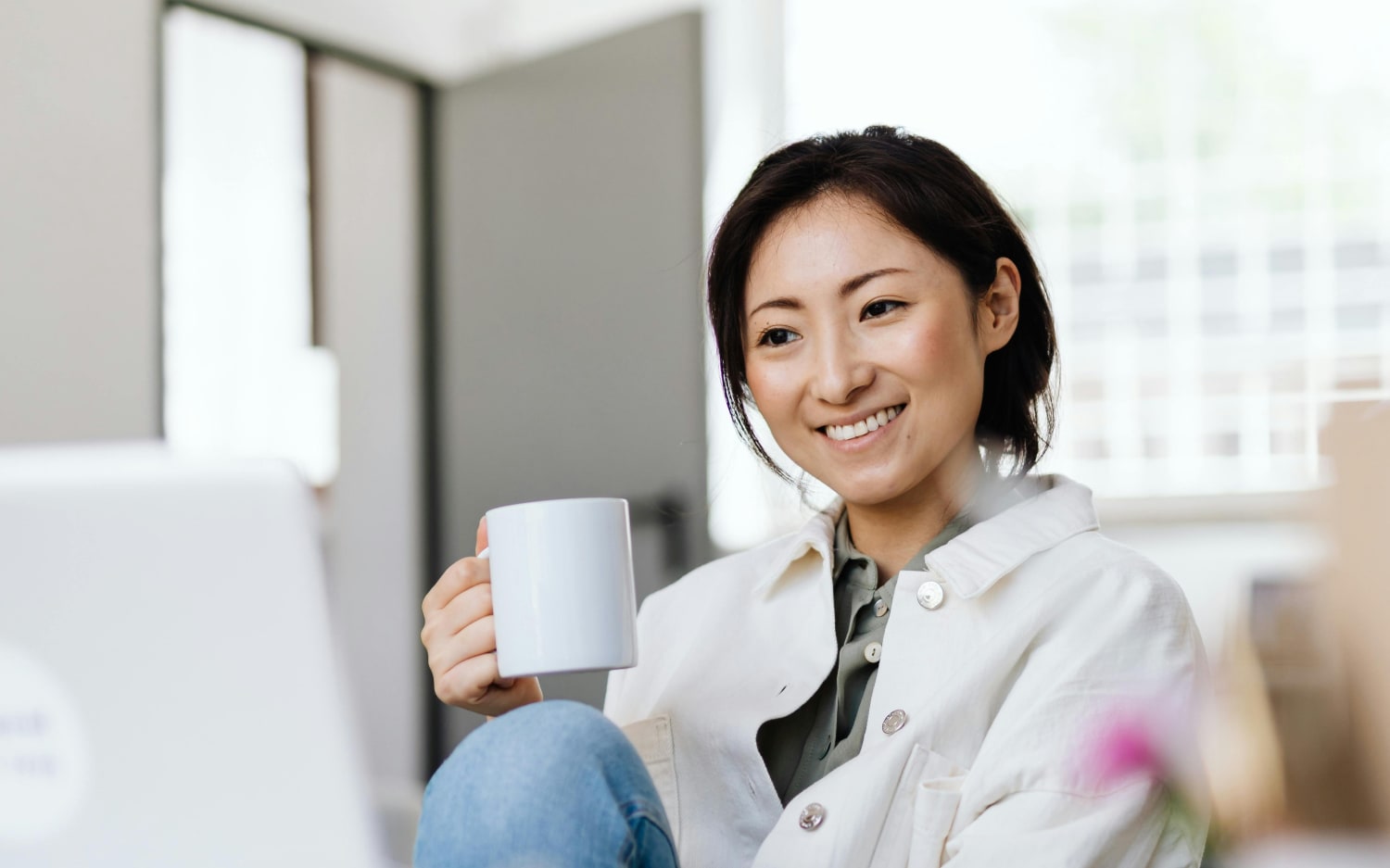 En smilende kvinne sitter i et lyst rom med en hvit kaffekopp i hånden og ser mot en bærbar datamaskin.