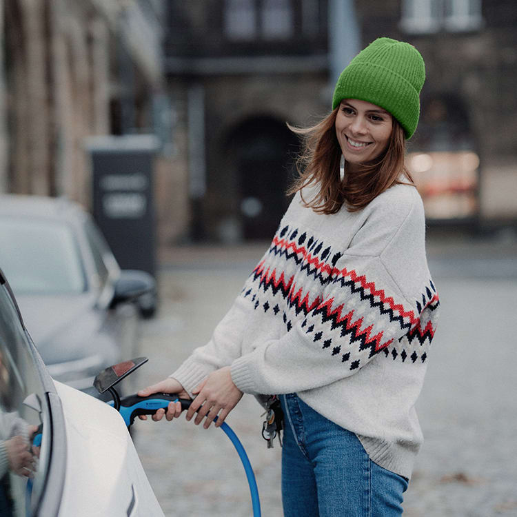Person lader en elbil ved en ladestasjon i byen og smiler mens hun holder ladekabelen.