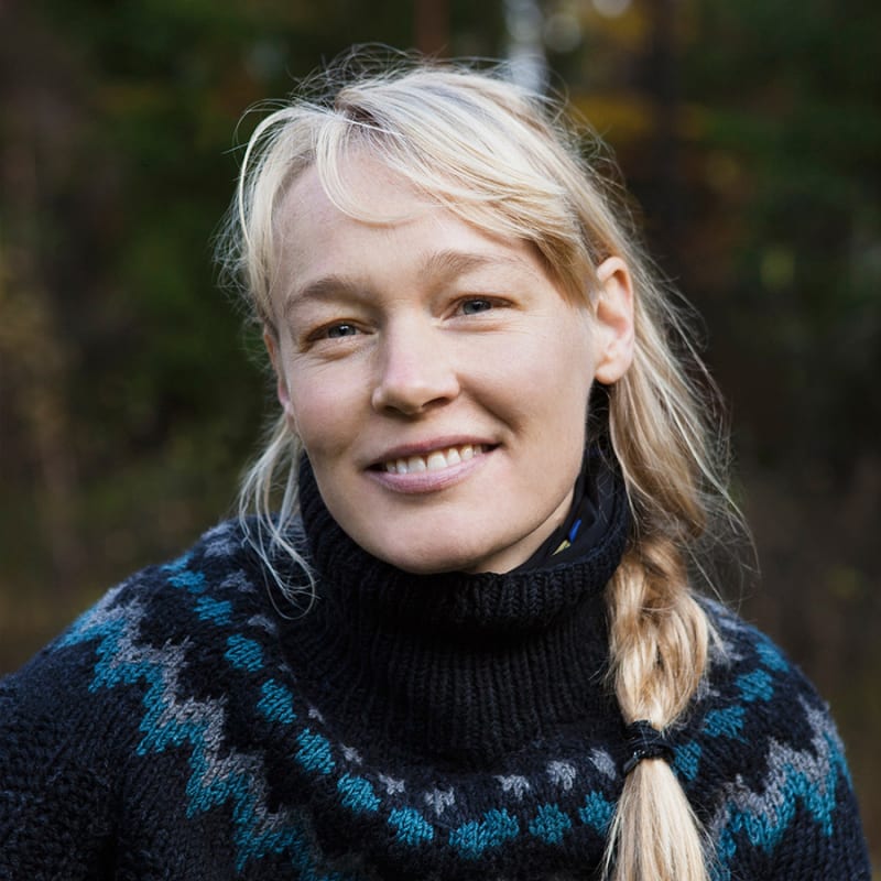 En blond kvinne med flettet hår og strikket genser smiler mot fotografen