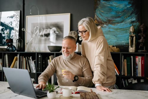 En mann og en kvinne sitter sammen ved et bord og ser smilende på en bærbar datamaskin, med en bokhylle og kunstverk i bakgrunnen.