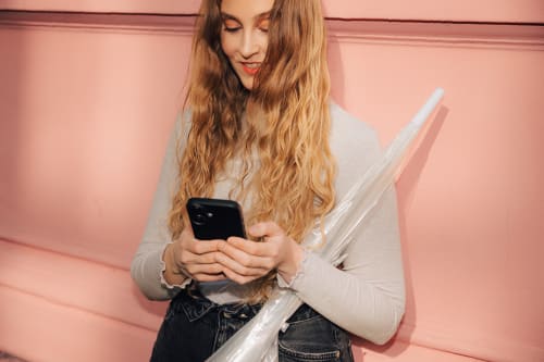 Betale med kredittkort i Vipps: ung kvinne ser på mobilen foran en rosa vegg