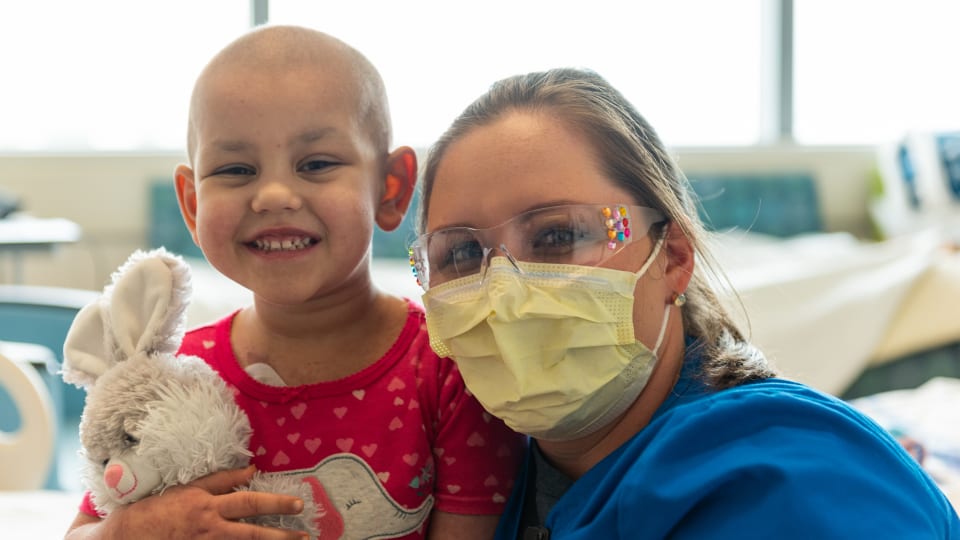 masked nurse poses with a smiling young patient holding a stuffed animal