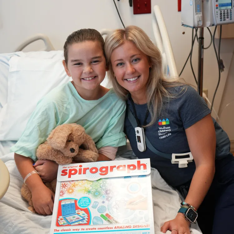 child life specialist next to a smiling pediatric patient in her hospital bed