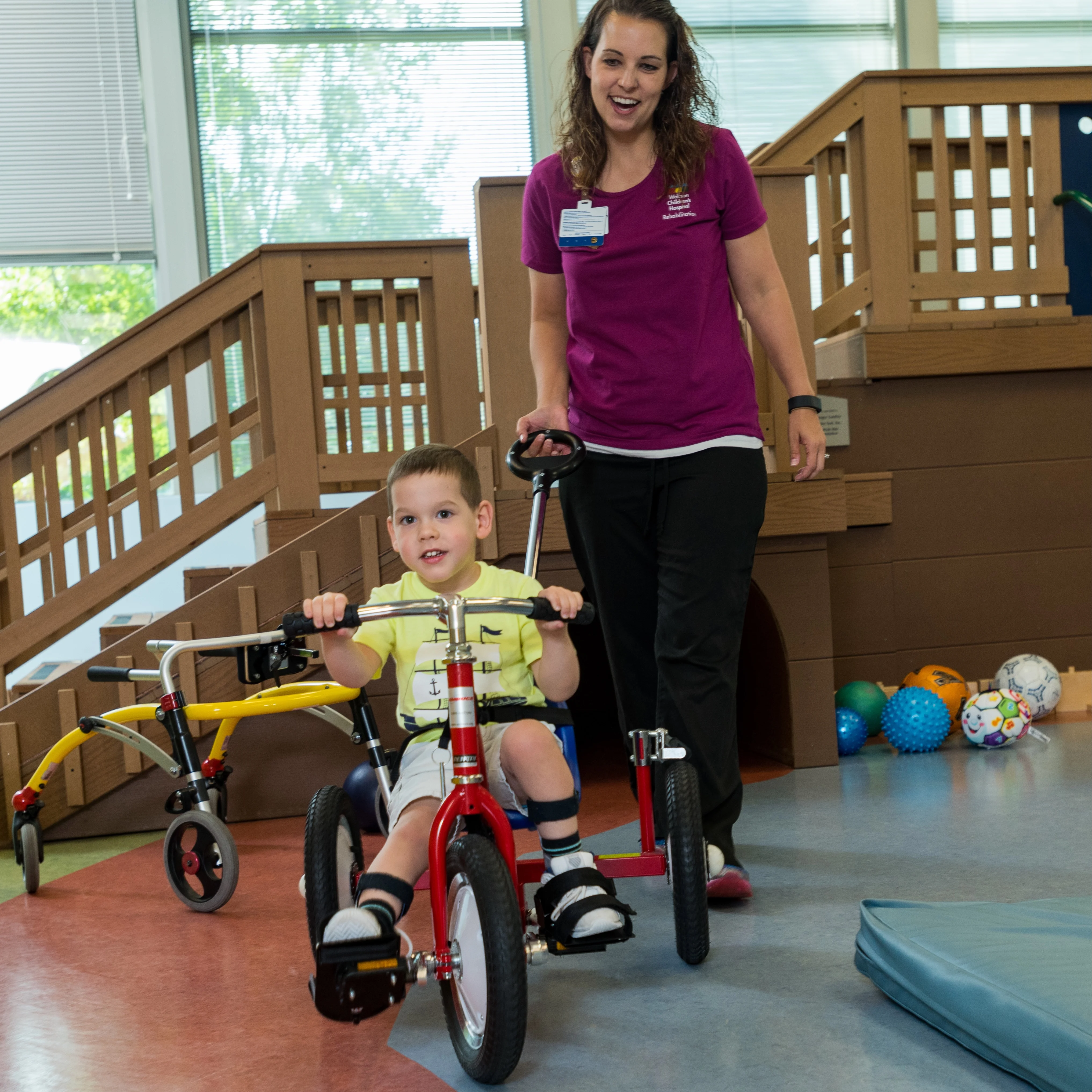 photo of a pediatric clinic setting where a female provider pushes a young male patient on a tricycle