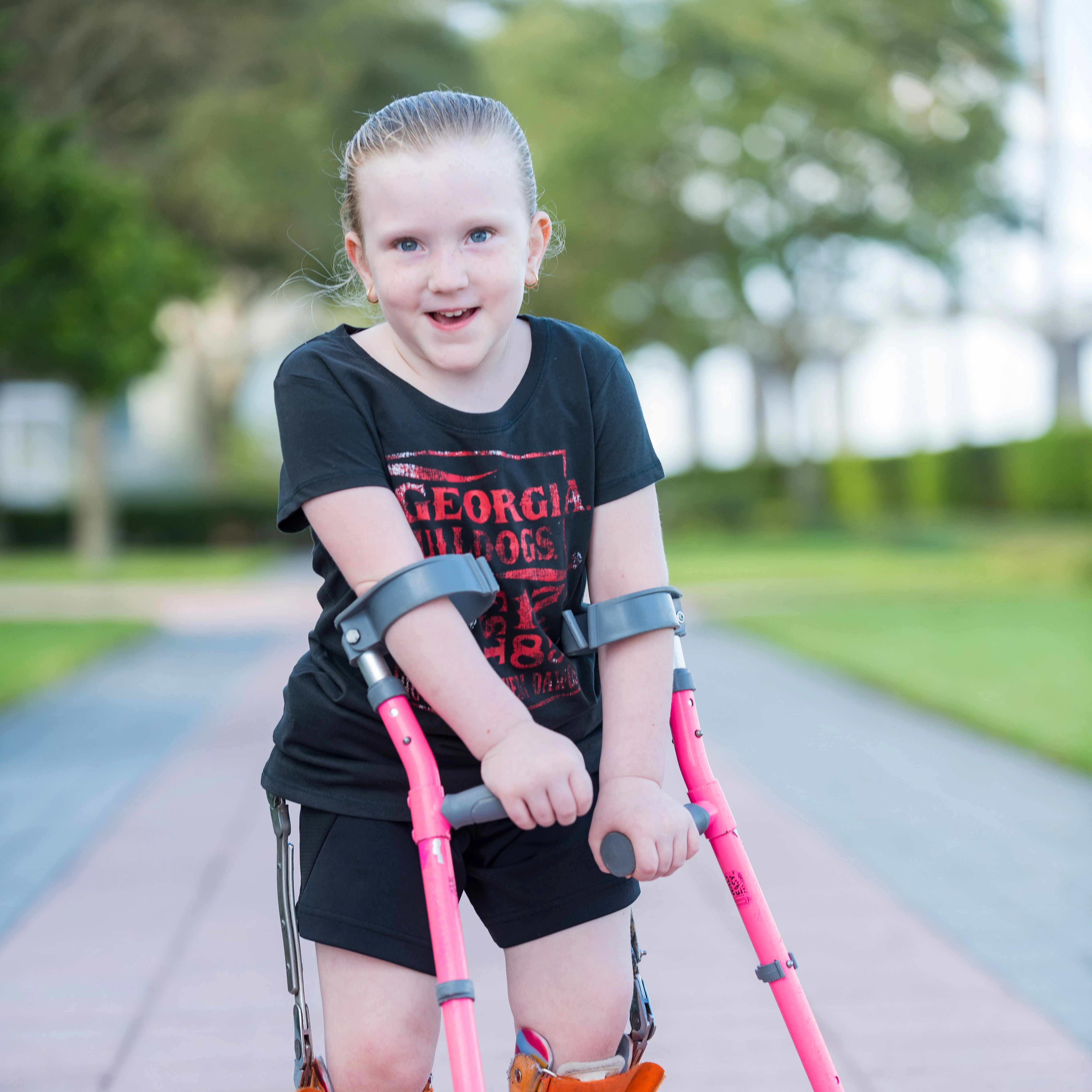 smiling young girl stands outside on the sidewalk using pink forearm crutches