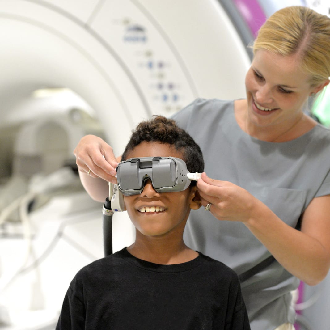woman helps put video goggles on a smiling young male patient to help relax him before undergoing MRI