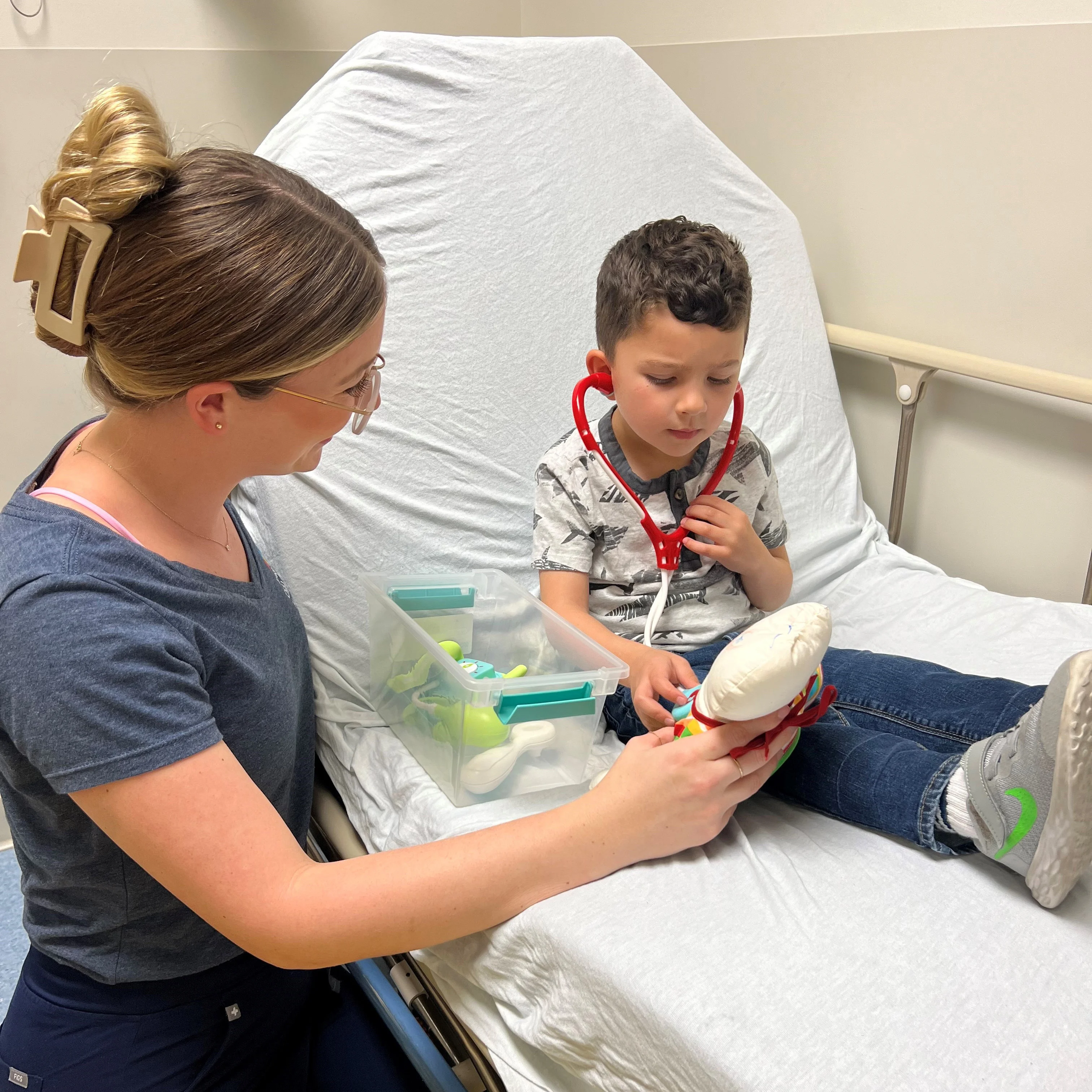 smiling female child life worker helps young boy in hospital bed understand a procedure by using a toy stethoscope and stuffed animal