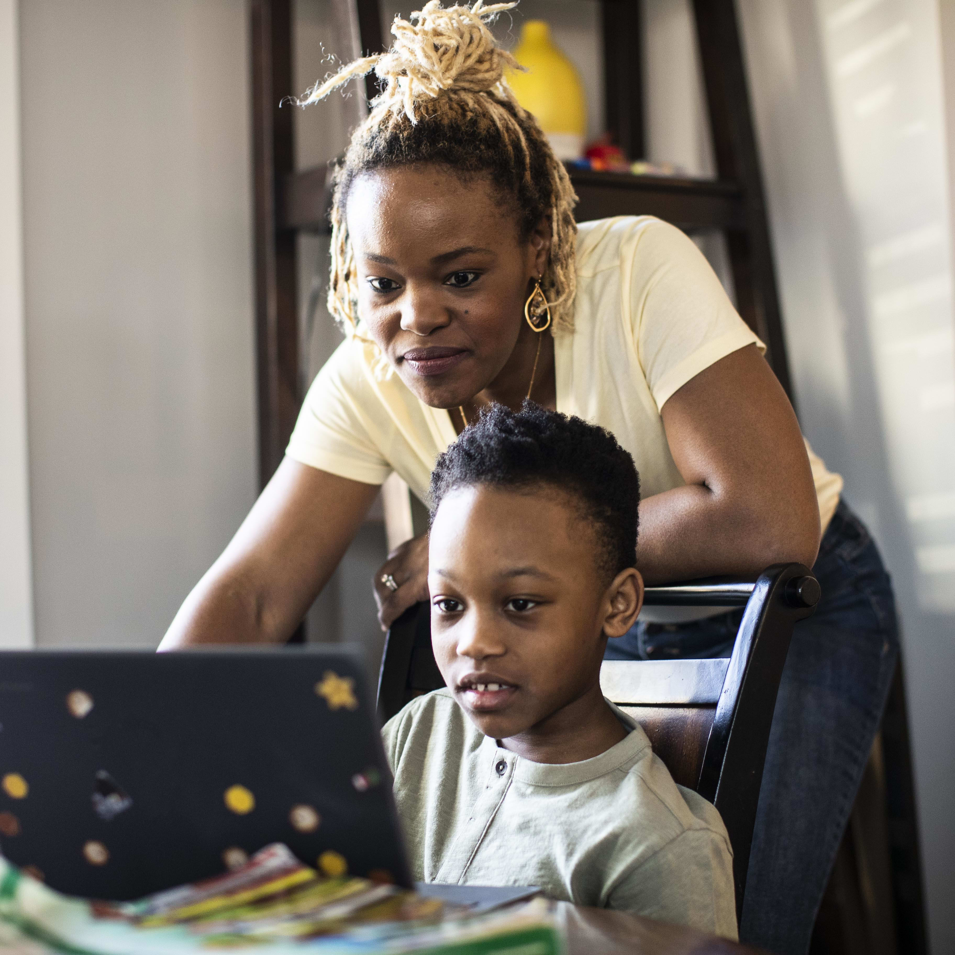 an African American mother looks over her son's shoulder as they both look at the screen of a laptop computer.