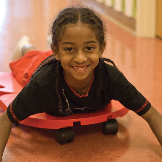 smiling child lays on red scooter boards in a hallway