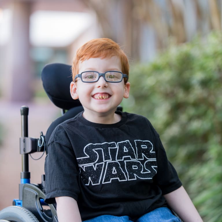 smiling elementary age boy wearing a Star Wars shirt and sitting in a wheelchair outside
