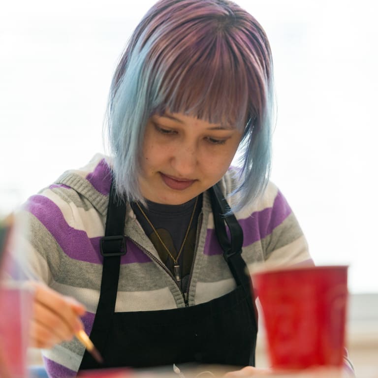 a female teenager with purple-and-blue-dyed hair wears a black smock while sitting at a table and drawing with colored pencils
