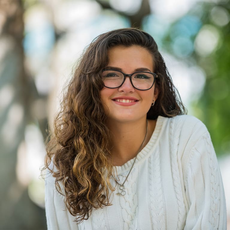 close up of a teenage girl in glasses and a sweater smiling at the camera while sitting outside in front of a tree