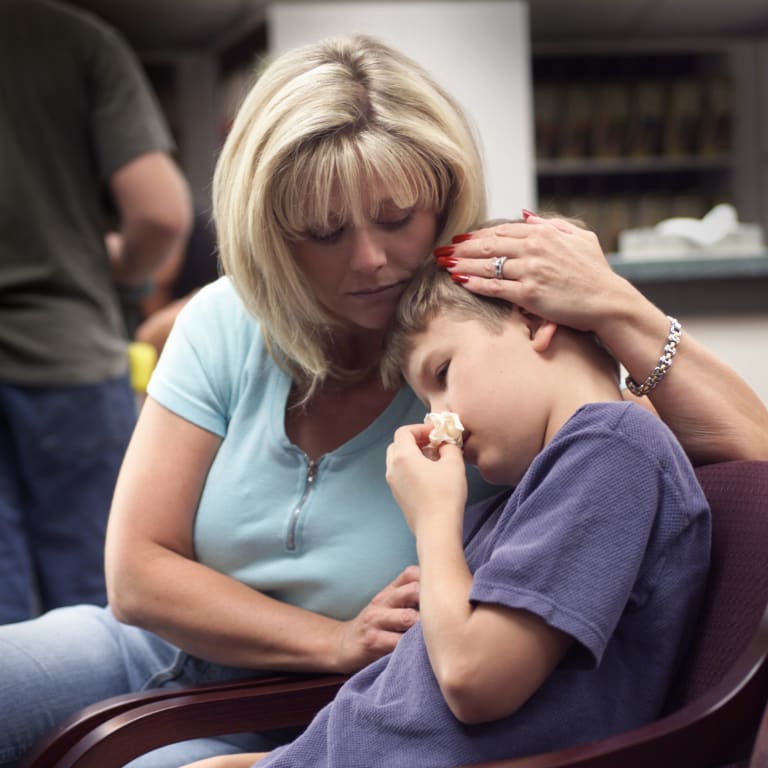 mother holder her elementary-aged son's head on her shoulder while he hold a tissue to his nose