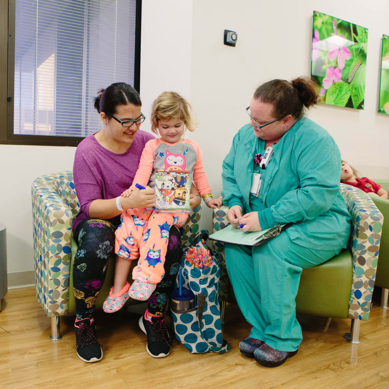 young girl sitting on her mom's lap looking at a book while a medical professional looks on