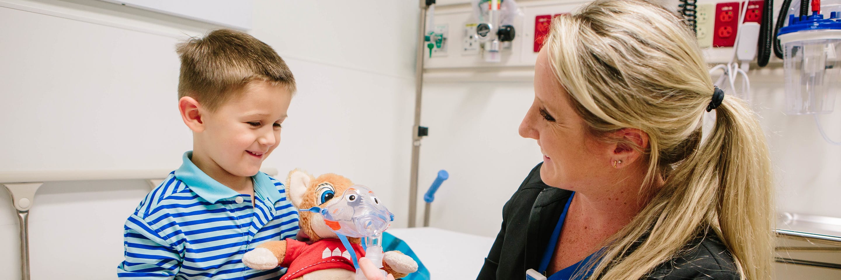 nurse hands a young boy in a hospital room a stuffed animal wearing a mask