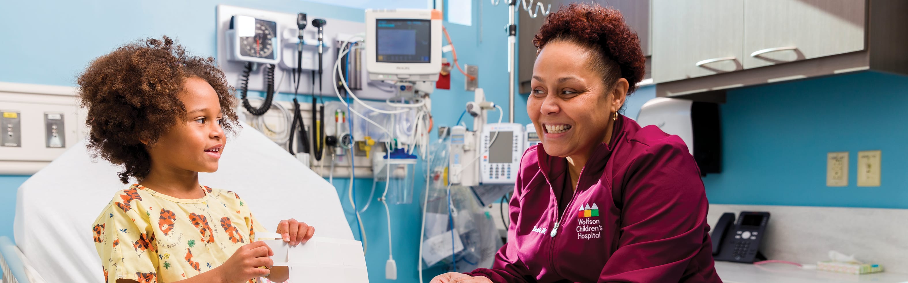 a smiling female nurse sits beside a smiling young girl in a hospital bed