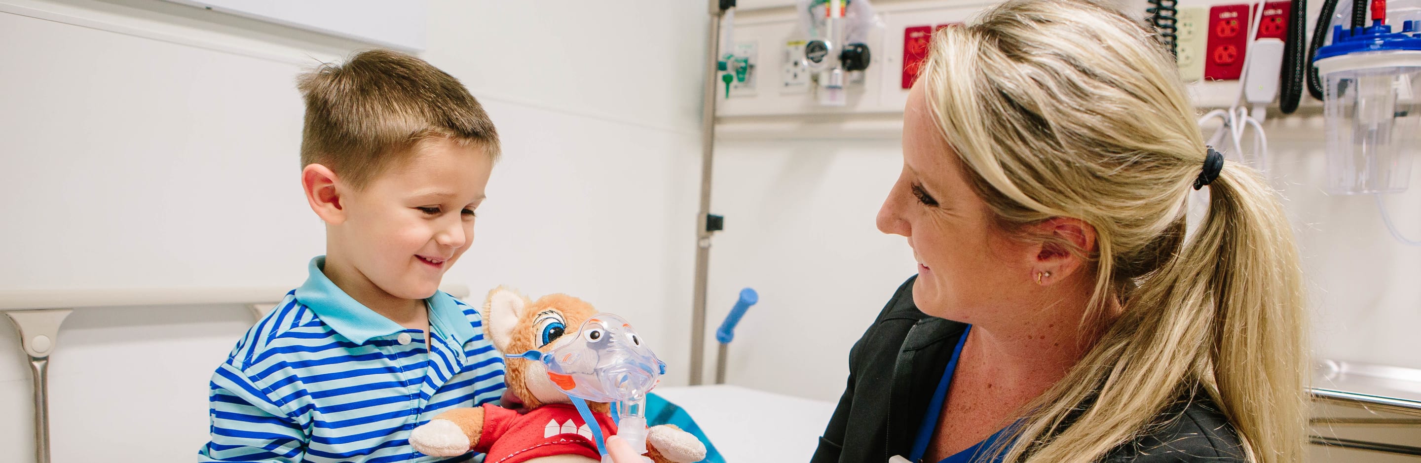 nurse hands a young boy in a hospital room a stuffed animal wearing a mask