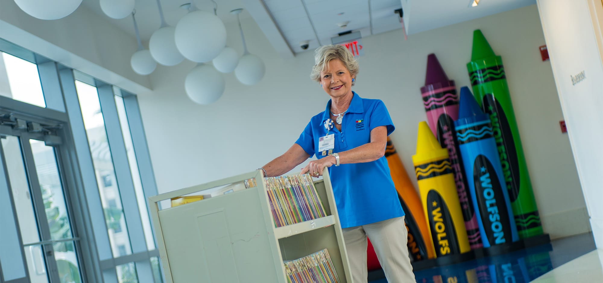 Smiling woman in blue Wolfson Children's shirt pushes a cart of books down a hospital hallway