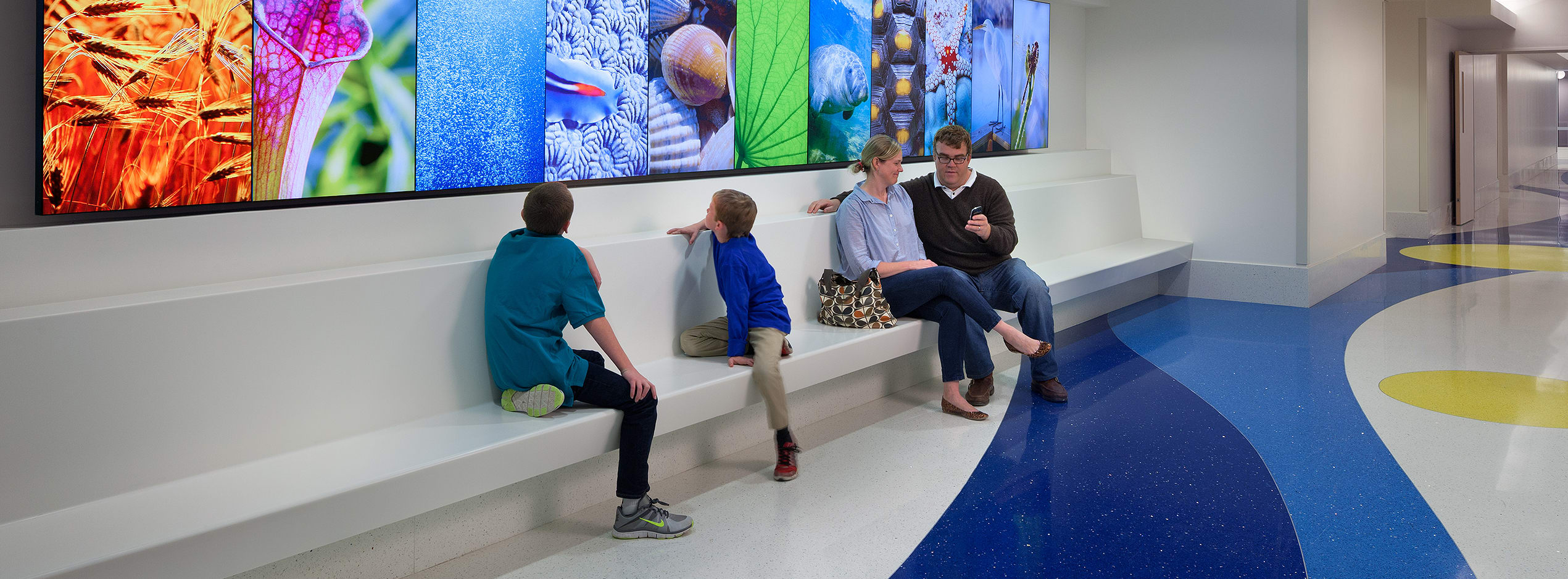 mom, dad and 2 young boys sitting on a bench in a brightly lit hospital hallway
