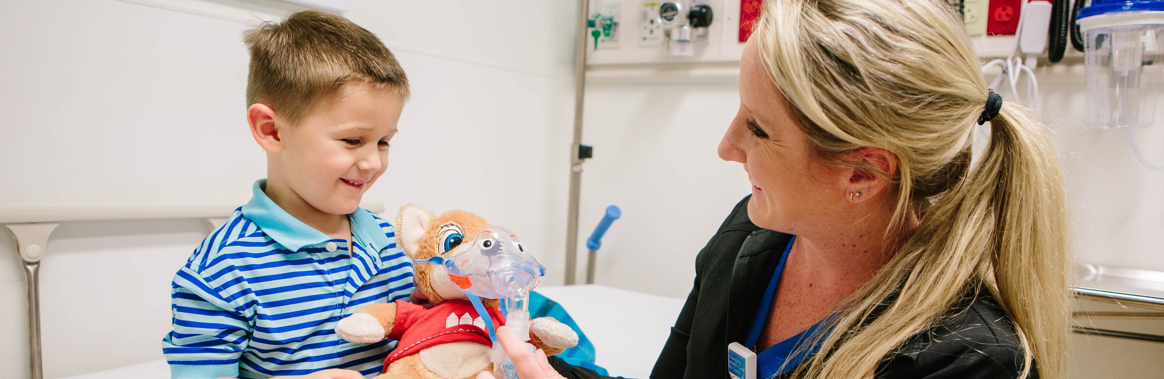 nurse hands a young boy in a hospital room a stuffed animal wearing a mask