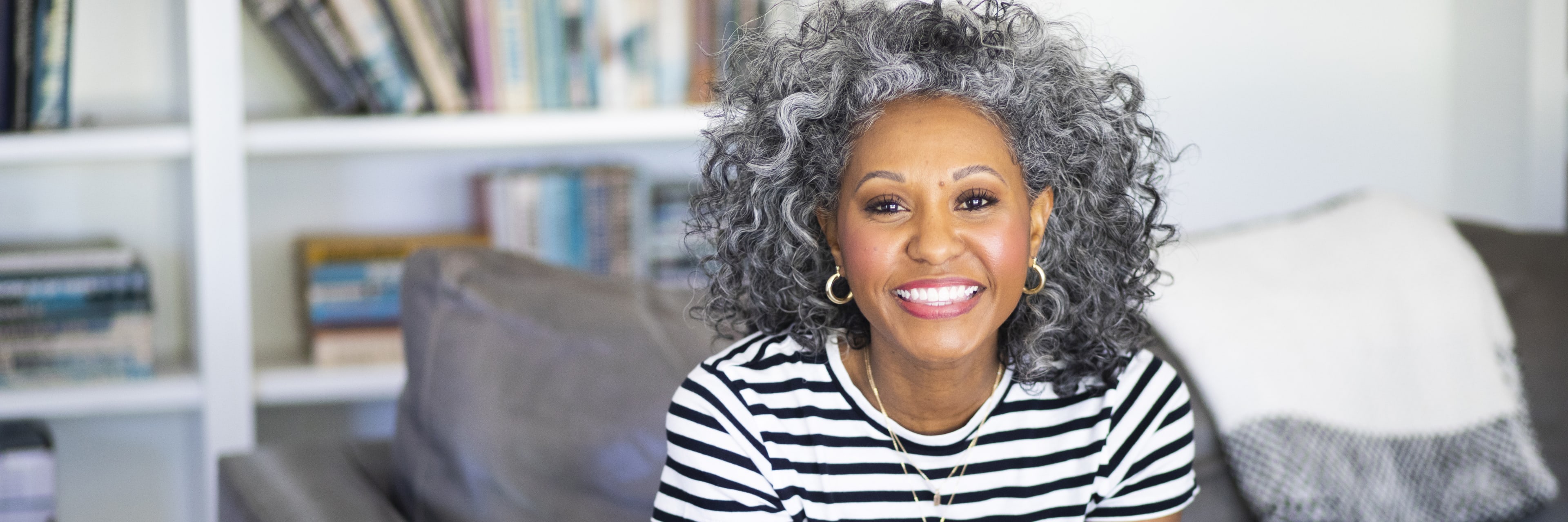 A middle-aged woman with curly hair sitting on a couch in front of a bookcase smiling