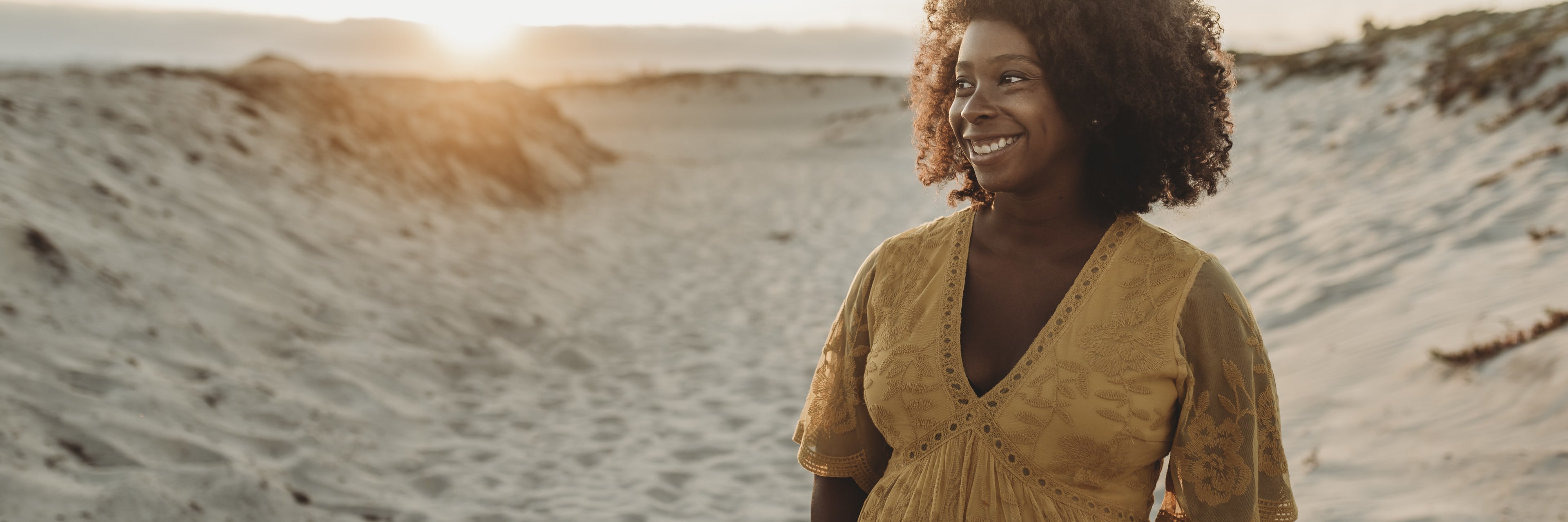 a pregnant woman looks happy cradling her baby bump while she stands in the midst of sand dunes at the oceanfront.
