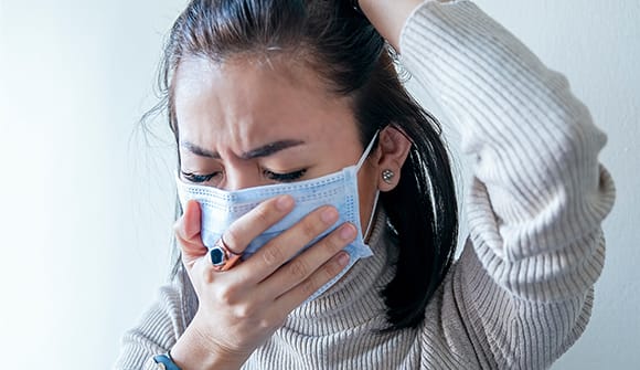 woman wearing a medical mask holds hand over her face while making a pained expression