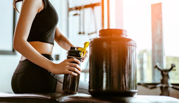 Women sitting on her kitchen counter holding a water bottle that she is loading protein powder into.