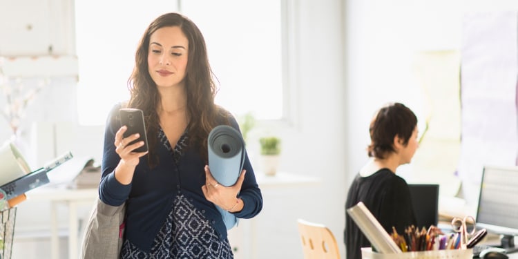 woman looking at her phone, holding a yoga mat in a brightly lit office space