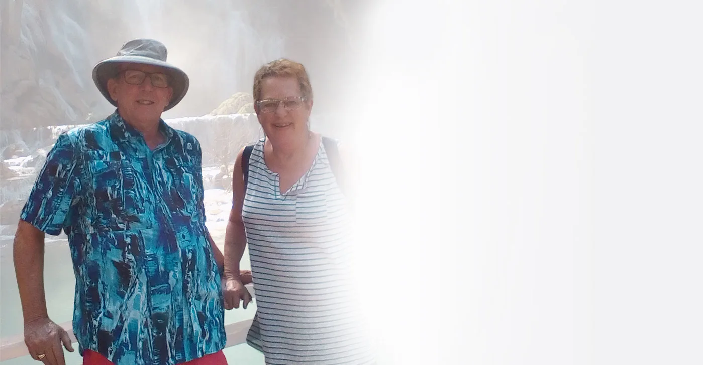 A middle-aged man and woman in vacation attire smile together in front of a waterfall.