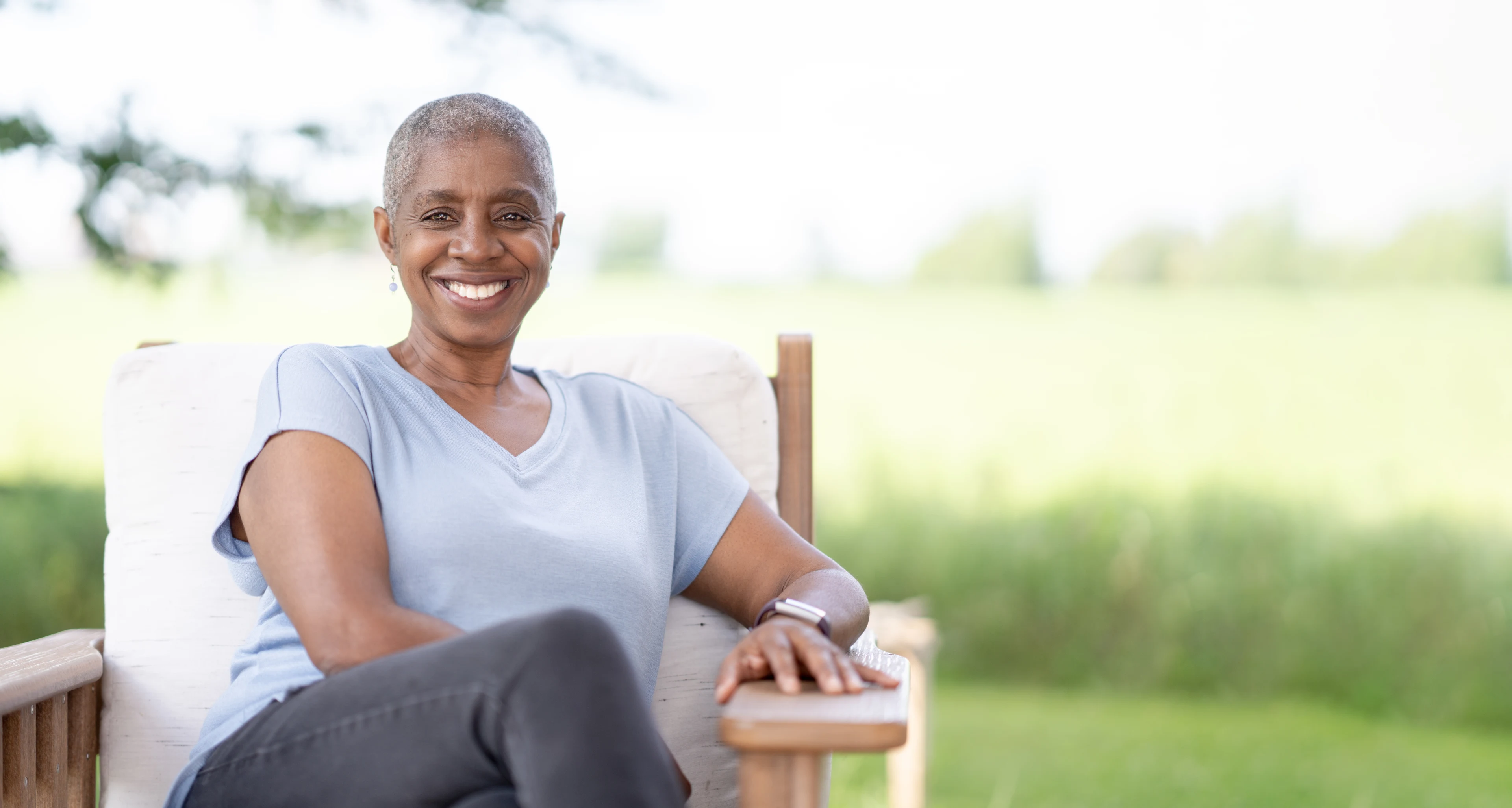 A middle aged woman sits outside on a sunny summer day dressed casually and smiling.