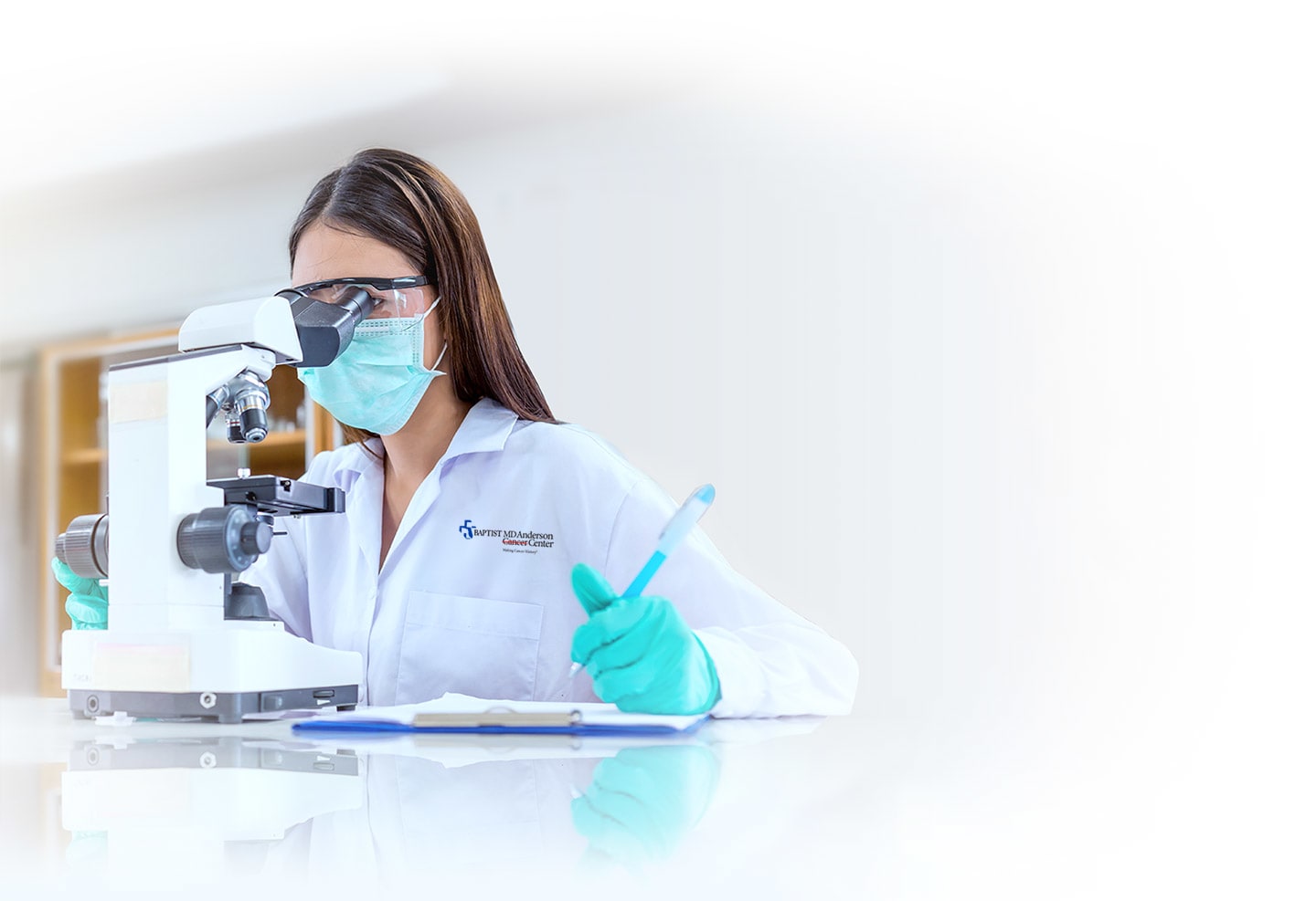 A female medical researcher wearing safety goggles, surgical mask and gloves, peers through a microscope, making notes on her clipboard.