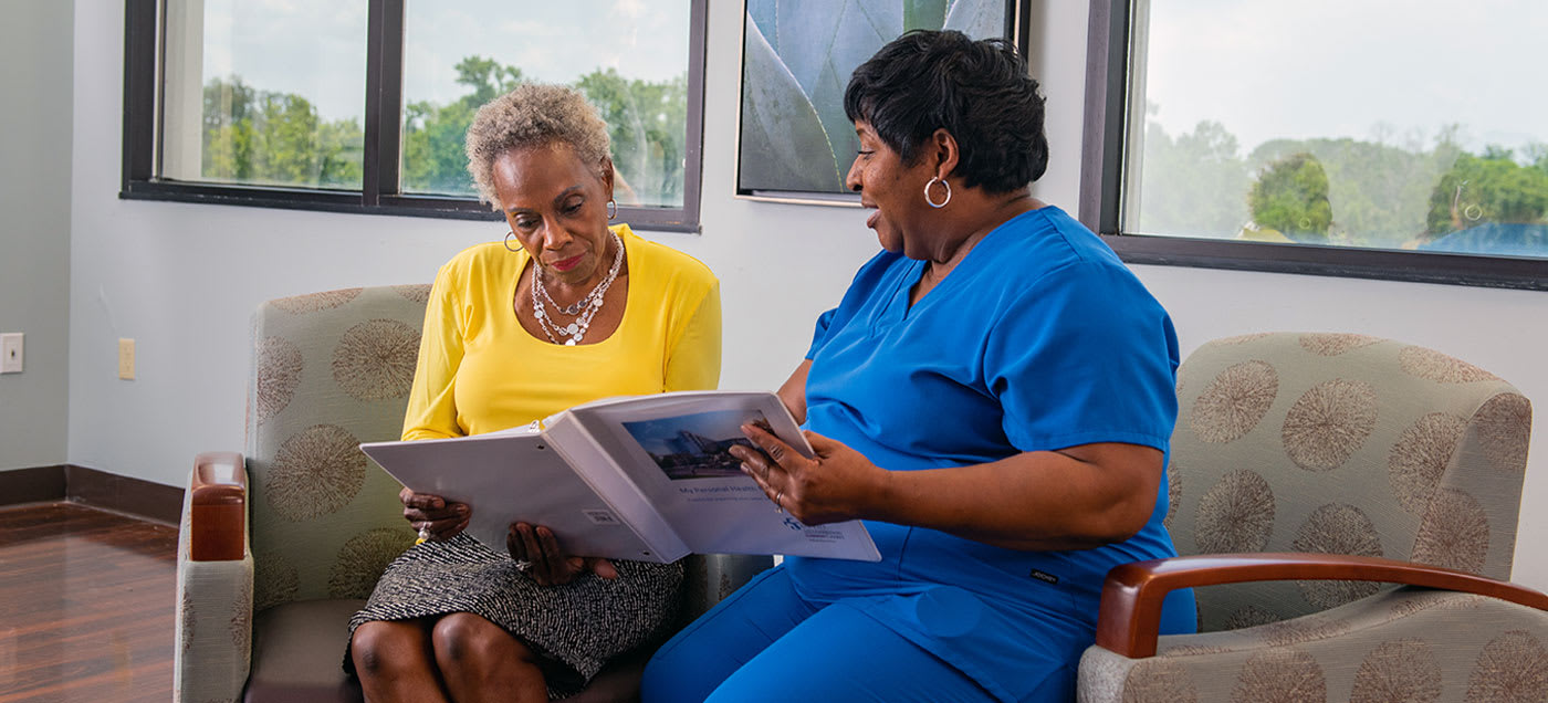 a nurse and a well-dressed woman sitting in chairs looking over a binder of patient resource information together