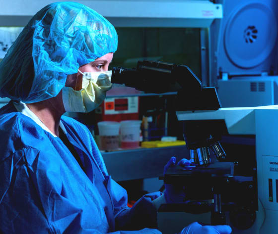 A closeup of a researcher's face, wearing mask and hair covering, looking through a microscope.