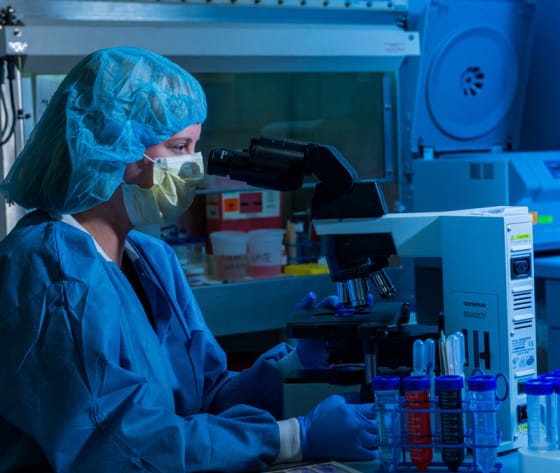 A closeup of a researcher's face, wearing mask and hair covering, looking through a microscope.