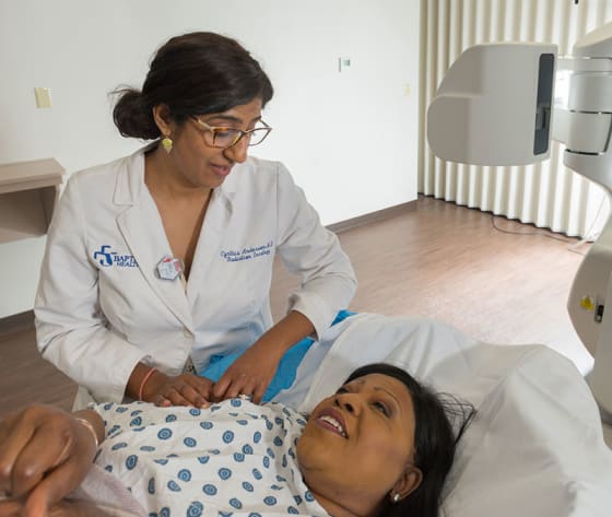 Dr. Cynthia Anderson sits, chatting with a female patient who lays in a gown, ready for radiation treatment.