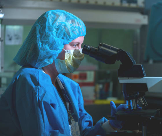 A researcher wearing gloves and mask looking through a microscope.
