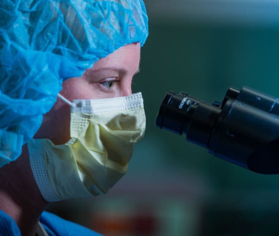 A closeup of a researcher's face, wearing mask and hair covering, looking through a microscope.
